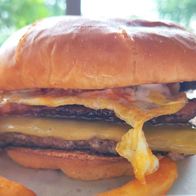 Close-up of a stacked burger featuring a soft bun, layers of melted cheese, crispy bacon, a fried egg, and a juicy beef patty, accompanied by curly fries, highlighting the culinary creativity of Portland's Burger Week.